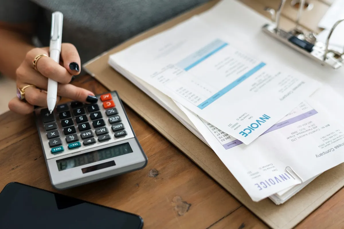 Mão com caneta usa uma calculadora ao lado de faturas impressas