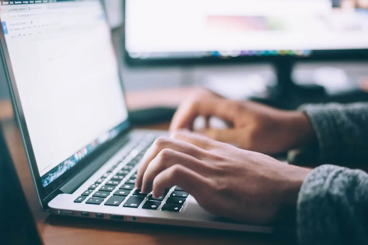 Mãos digitam em um notebook diante de um monitor