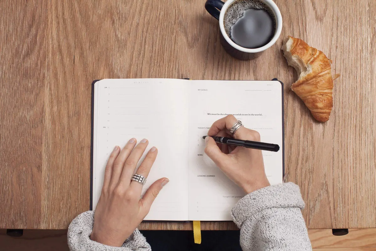 Mão escreve em um caderno ao lado de uma xícara de café e um croissant