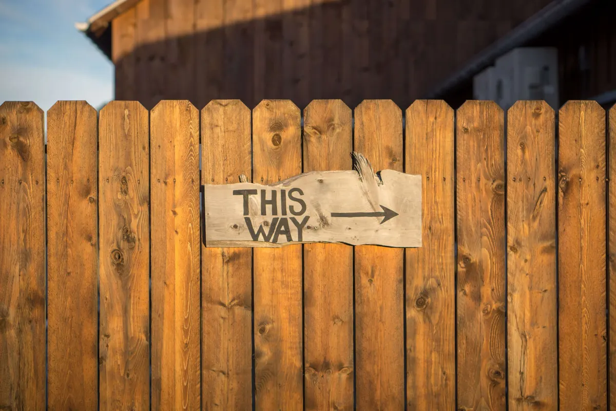 Placa de madeira com a inscrição This way e uma seta, em uma cerca